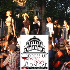 Line of models on a catwalk parading behind Dress Up Wine Down on Cap logo, May Second Saturday.
