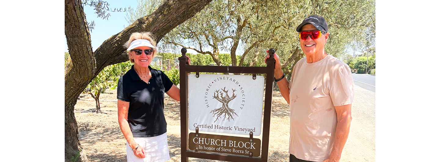 Two people standing with their hands on a sign reading Church Block in front of olive trees and vines