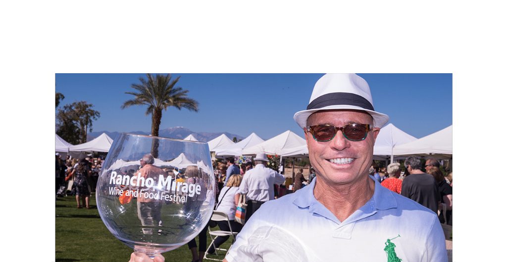 Very happy smiling man holding glass of red wine with Rancho Mirage logo.